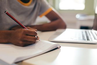 Close-up of a student taking notes with a pencil and a laptop on a desk.