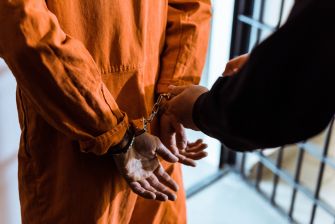 A person in an orange jail jumpsuit is in handcuffs, with another person standing nearby.