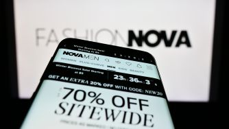 Close-up of a smartphone displaying a Fashion Nova promotional banner, with the Fashion Nova logo visible on a blurred screen in the background.