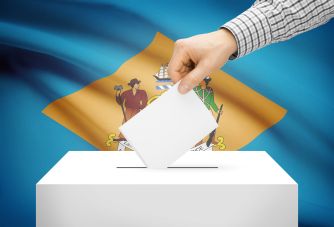 Close-up of a hand dropping a ballot into a ballot box with the Delaware state flag in the background, representing voting in Delaware elections.