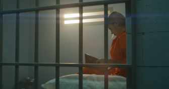 Prison inmate in an orange jumpsuit sits in a jail cell, reading a folder.