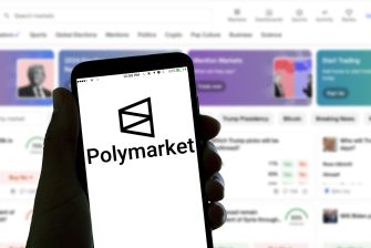 A hand holds a smartphone displaying the Polymarket logo, with a blurred webpage in the background.