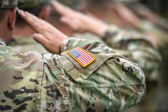 Soldiers in camouflage uniforms saluting, with a U.S. flag patch on their sleeve.
