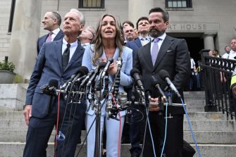 A woman in a light-blue suit speaks at a podium surrounded by multiple microphones, flanked by men in suits on courthouse steps.