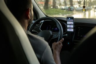 Person driving a car with a smartphone mounted on the dashboard displaying a rideshare app.