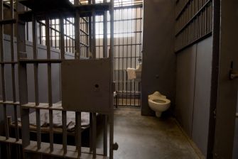 Prison cell interior with metal bars, a bare bed frame, and a toilet.