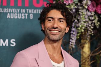 Smiling man with dark hair wearing a light pink suit at a formal event, standing in front of a floral backdrop.