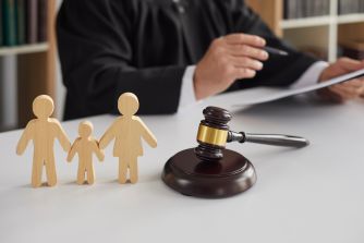 Gavel and wooden family figures on a desk with a person signing documents in the background, symbolizing a child-welfare settlement.