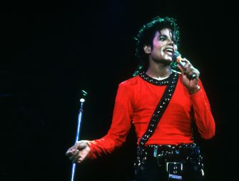 Singer on stage wearing a red shirt and studded belt, holding a microphone during performance.
