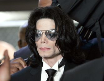 Person with long dark hair wearing a black suit, white shirt, black tie, and large sunglasses.