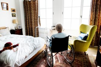 Elderly resident in a wheelchair facing a large window in a bright nursing home room with a bed and a yellow armchair nearby.