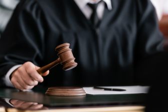 Close-up of a judge's gavel striking a sound block on a wooden desk, with a blurred figure in a dark suit in the background.