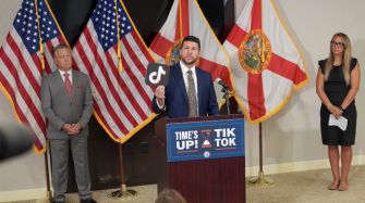 Press conference scene with a man speaking at a podium displaying a TikTok logo, flanked by a man and a woman, with American and Florida flags in the background.