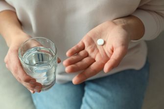 Close-up of a person’s hands, one holding a single white pill and the other holding a glass of water.