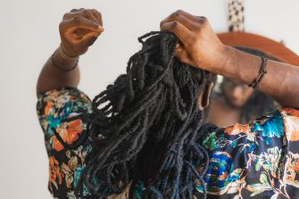 Person with long dreadlocks wearing a colorful shirt, with hands on their head while another person touches their hair.