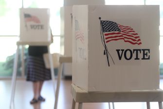 White ballot drop box stamped with VOTE and a US flag, indoors, with a blurred person in the background.