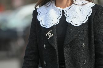 Person wearing a black coat with a white scalloped lace collar and a Chanel interlocking CC logo on the lapel.