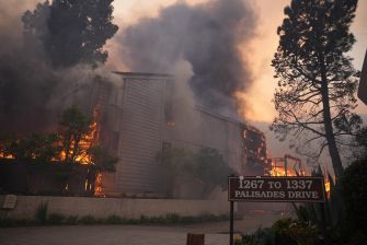 A house-like building engulfed in flames with thick smoke rising, surrounded by trees, and a street sign reading 1267 to 1337 Palisades Drive in the foreground.