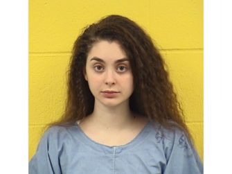 Mugshot-style portrait of a young woman with long dark hair, wearing a light blue shirt, standing against a yellow backdrop.