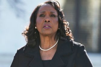 Portrait of a Black woman in a black blazer and pearl necklace, outdoors, speaking.