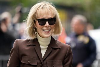 Smiling woman with blond hair and dark sunglasses wearing a brown coat at an outdoor event.