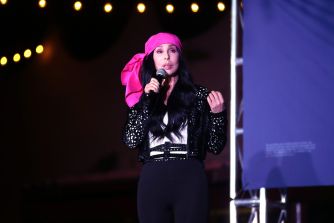 Female performer on stage wearing a pink headscarf and black sequined outfit, singing into a microphone under stage lights.
