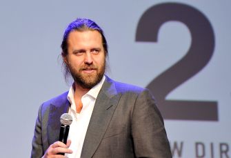 Bearded man in a gray suit holding a microphone and speaking on stage, with a large “2” on the screen behind him.