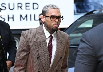 Man with short gray hair and glasses wearing a brown plaid suit, white shirt, and burgundy tie, standing outdoors next to a car.