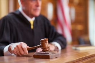 Gavel resting on a wooden judge's bench in a courtroom, with a judge blurred in the background.