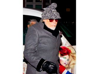Older man in a gray coat and knit hat with sunglasses and gloves, standing beside a car with a poster of a woman visible behind him.