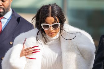 Woman wearing a white fur coat and white sunglasses walks outdoors, showing red nail polish.