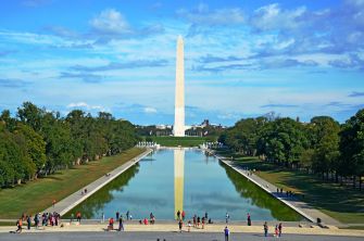 View of the Washington Monument reflected in the Reflecting Pool on the National Mall, with trees along the sides and people walking near the water.