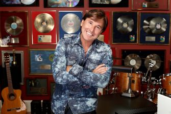 Smiling man in a denim shirt stands in a music studio, surrounded by gold record plaques and musical equipment.