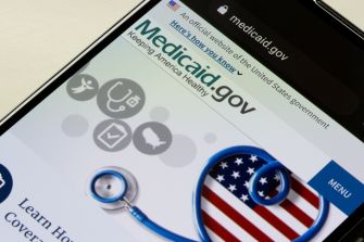 Close-up of a smartphone displaying the Medicaid.gov homepage with medical icons and a stethoscope beside a U.S. flag motif.
