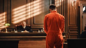 A man in an orange prison jumpsuit stands in a courtroom facing the judge at the bench.