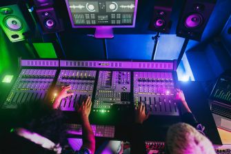 Overhead view of a music recording studio with a large mixing console, hands on the faders, and colorful ambient lighting.
