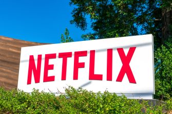 Netflix logo on a white signboard outdoors, with green bushes and a blue sky in the background.