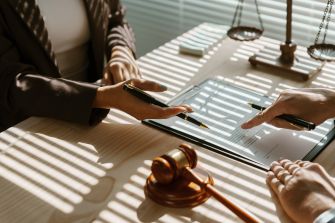 Close-up of two people’s hands reviewing legal documents at a desk, with a gavel and sunlight streaming through blinds.