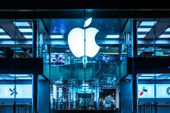 Exterior view of an Apple Store with a large glowing Apple logo inside a blue-lit storefront.