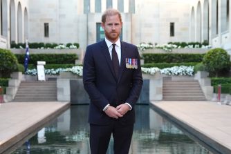 Man with red hair in a dark suit wearing medals, standing with hands clasped in front at an outdoor formal setting with steps and a reflecting pool.