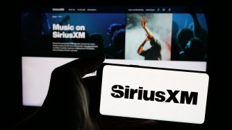 Close-up of a hand holding a white card with the SiriusXM logo, in front of a monitor displaying SiriusXM content.