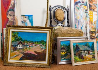 Collection of framed paintings leaning against a wall indoors, including landscapes and a portrait, with a decorative chair in the background.