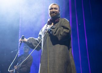 Male performer with a beard on stage singing into a microphone, wearing a long brown robe, with a guitarist in the background and blue stage lighting.