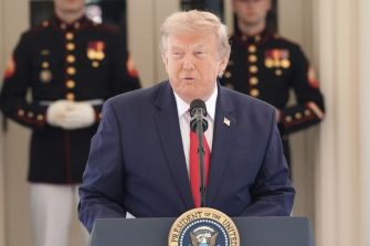 A man in a dark suit and red tie speaks at a podium bearing the presidential seal, with uniformed guards in the background.
