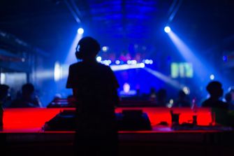 Silhouette of a DJ standing at a booth in a dim nightclub with blue and purple stage lights.