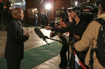 Man in a dark suit being interviewed by reporters with microphones and cameras outside a venue at night.