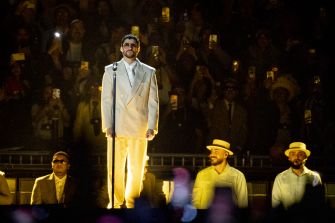 Male performer in a pale suit and sunglasses center stage with a microphone, flanked by two backup performers in white outfits and hats, audience visible in the background.