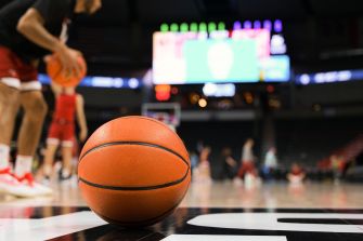 Close-up of a basketball on a court with players and a scoreboard blurred in the background.