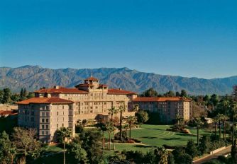 Resort-style hotel complex with beige walls and red-tiled roofs, surrounded by palm trees, set against a mountainous backdrop.