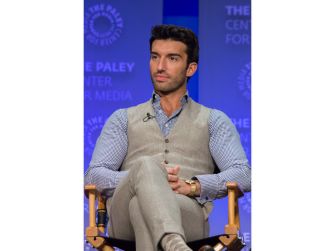 Man seated on a wooden chair on stage, wearing a light blue shirt and gray vest.